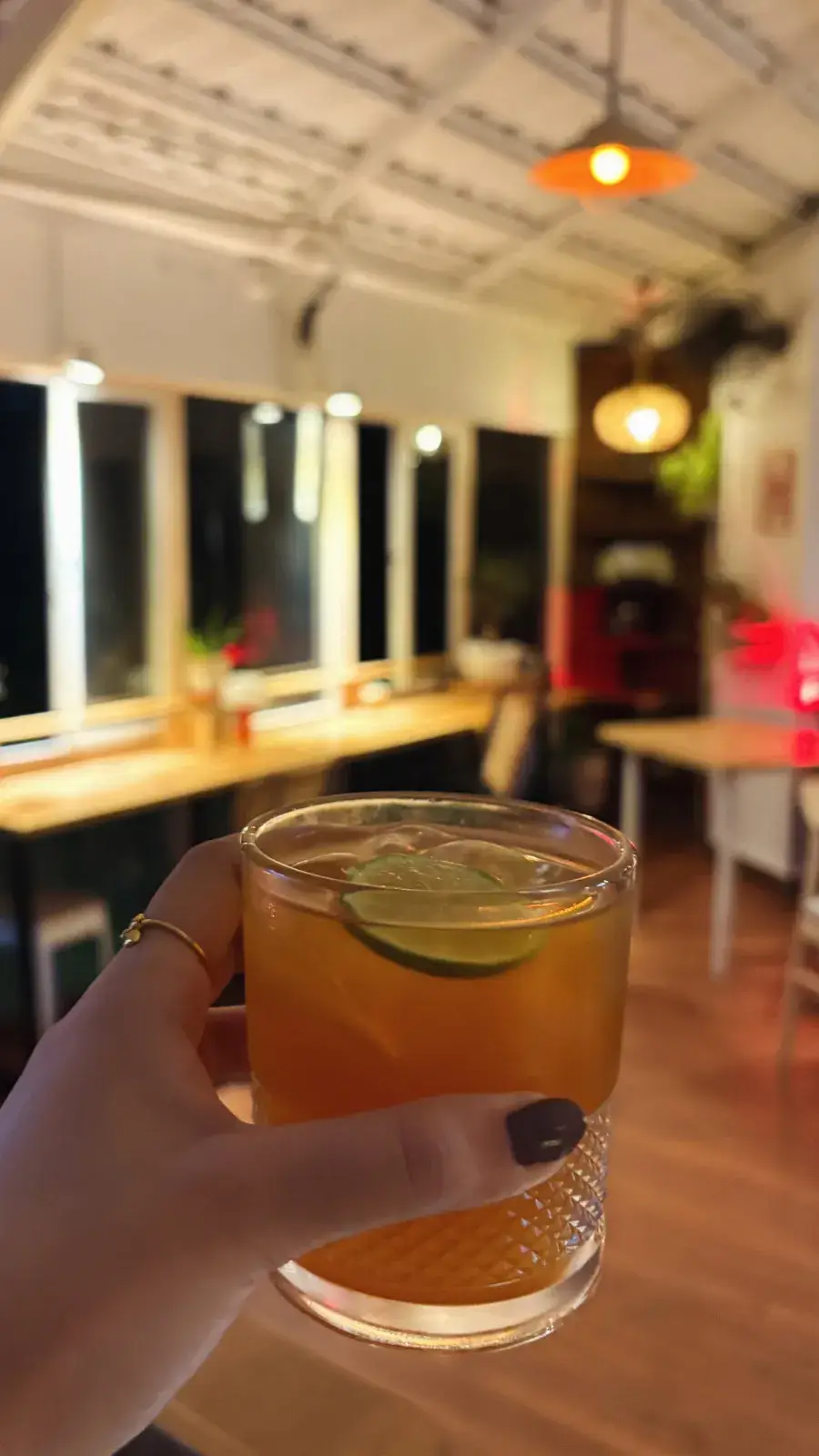 A lime cocktail held up in the lamp-lit room at night, the window counter and pendant lights behind