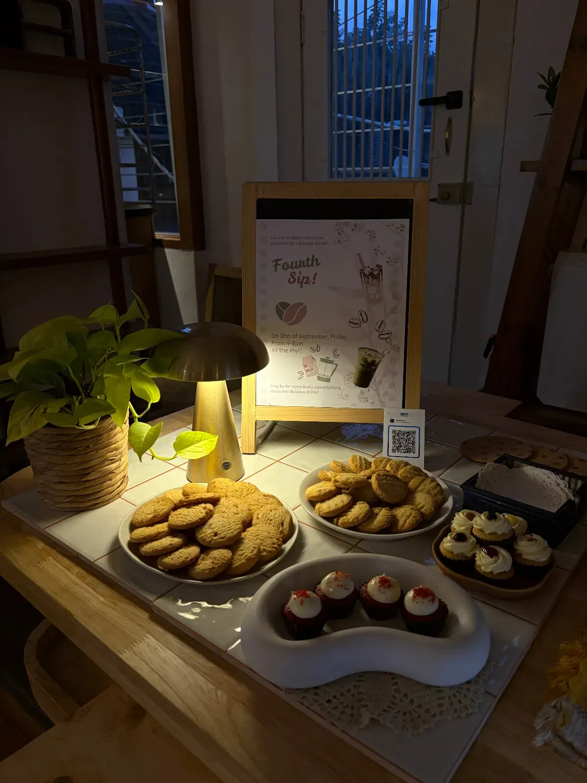 The pop-up table at dusk, cookies and red velvet cupcakes glowing under a brass lamp
