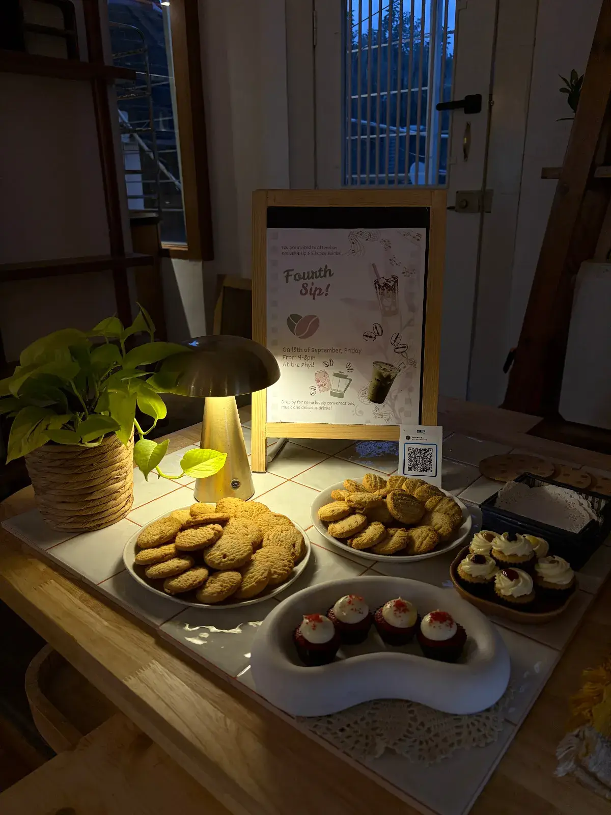 The pop-up table at dusk, cookies and red velvet cupcakes glowing under a brass lamp