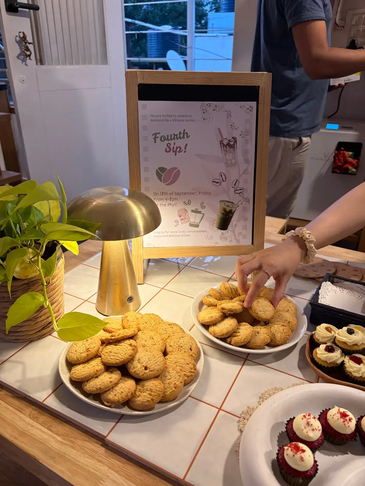 A Sip and Glimpse pop-up table: plates of cookies, cupcakes, a brass mushroom lamp and the event poster