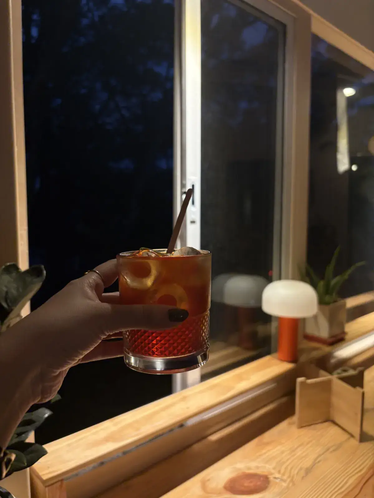 A cocktail raised at the window counter after dark, a small mushroom lamp glowing on the sill